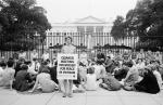 A group of people seated on the sidewalk outside the iron fence at the White House. One woman is standing, holding a sign that reads "Quaker Meeting for worship for peace in Vietnam."